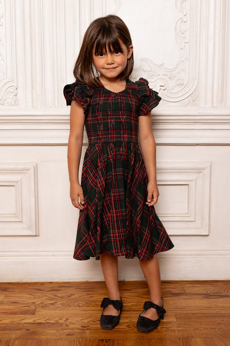 Young girl wearing a black and red plaid dress standing against a white wall.