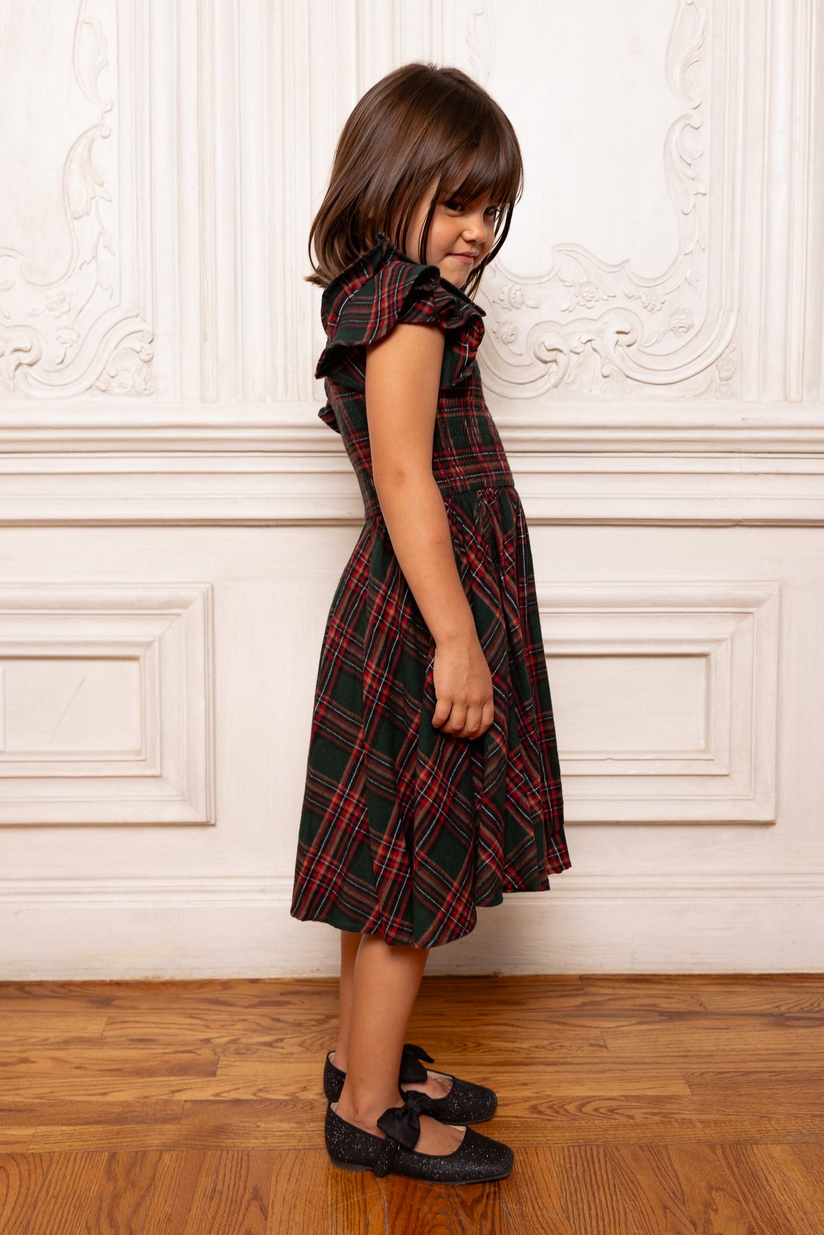 Young girl wearing a red plaid dress standing against a decorative white wall.