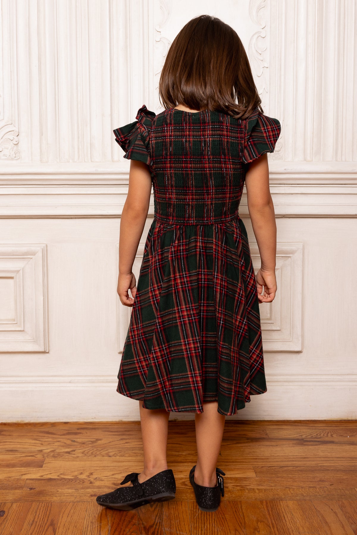 Child wearing a plaid dress standing against a white paneled wall.