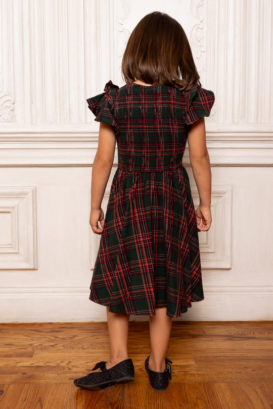 Child wearing a plaid dress standing against a white paneled wall.