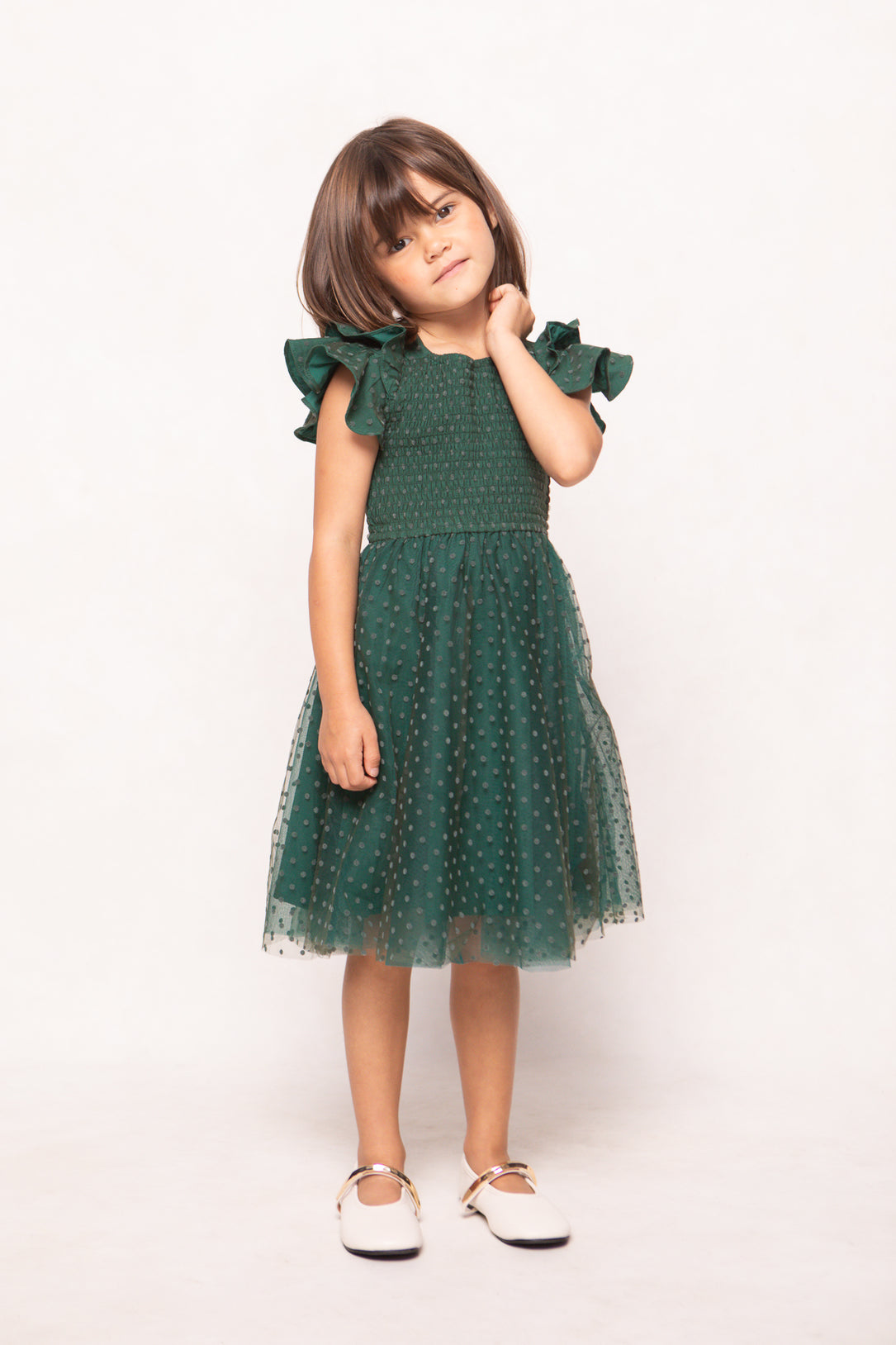 A young girl with straight brown hair wears the Ivy City Co Mini Hattie Dress in Green Flocked Dot Tulle, standing against a plain light background with her hand touching her neck. Perfect for mommy-and-me moments, she completes the look with white shoes.