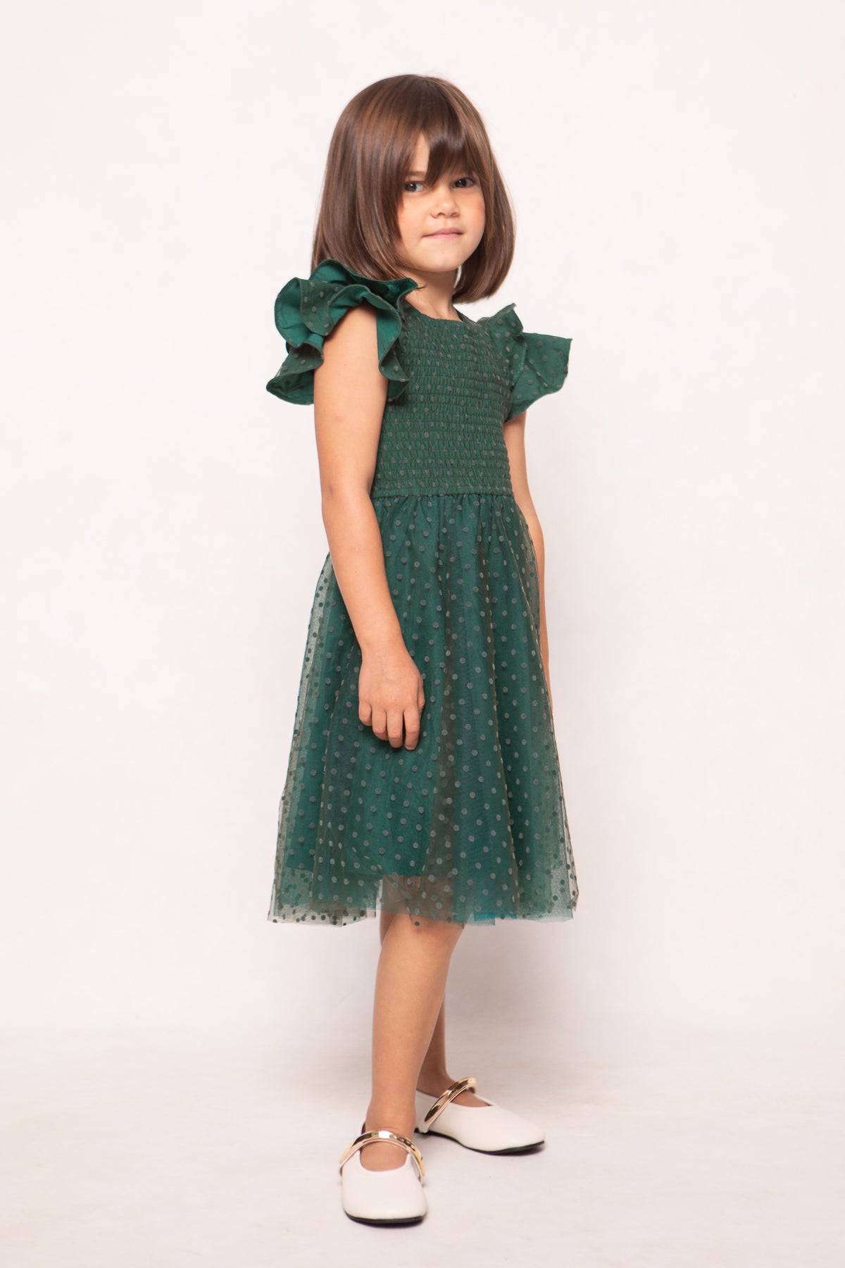 A young girl with brown hair stands against a plain, light background wearing the Ivy City Co Mini Hattie Dress in Green Flocked Dot Tulle—a short-sleeved, knee-length dress featuring a green flocked dot tulle overlay—paired with white shoes.