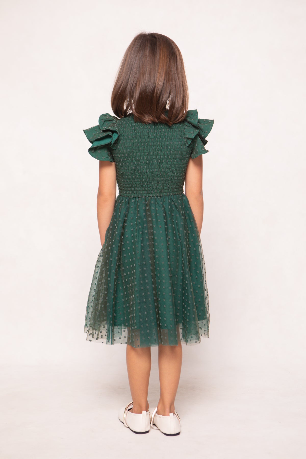 A young girl with shoulder-length brown hair stands facing away, wearing the Ivy City Co Mini Hattie Dress in Green Flocked Dot Tulle, which features ruffled sleeves and delicate green flocked dot tulle detailing. The knee-length dress is paired with white shoes, while the plain, light-colored background keeps the focus on her charming outfit.