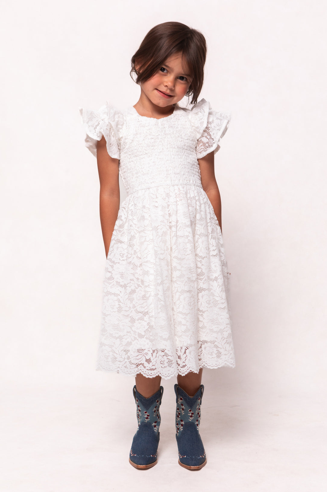 A young girl with brown hair in pigtails smiles while wearing the Ivy City Co Mini Hattie Dress in White Lace and blue cowboy boots, standing against a plain white background.