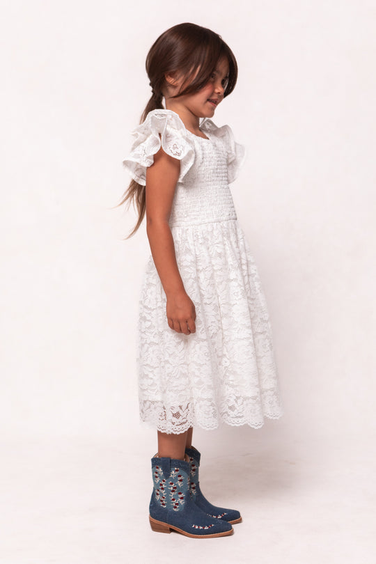A young girl with long brown hair smiles sideways in the Ivy City Co Mini Hattie Dress in White Lace, featuring ruffled sleeves, paired with blue embroidered cowboy boots, against a plain light background.