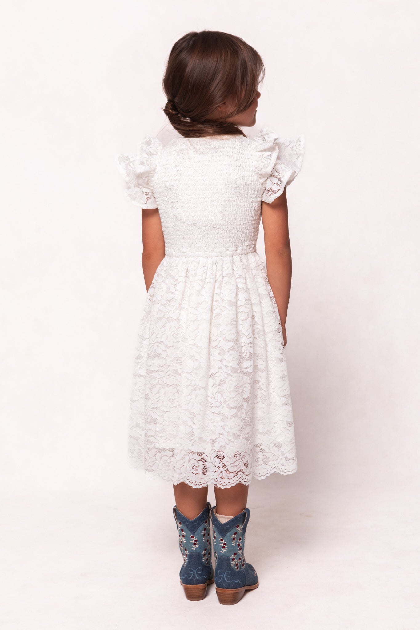 A young girl with brown hair, facing away, wears the Ivy City Co Mini Hattie Dress in White Lace paired with blue cowboy boots against a plain light background.