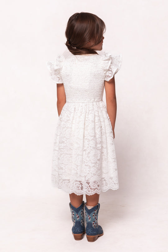 A young girl with brown hair, facing away, wears the Ivy City Co Mini Hattie Dress in White Lace paired with blue cowboy boots against a plain light background.