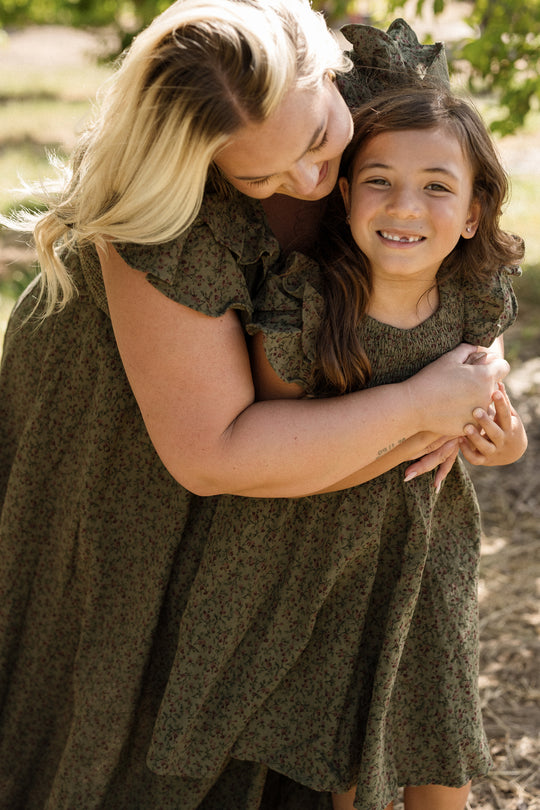 A woman hugs a smiling young girl outdoors, both wearing the Ivy City Co Mini Hattie Dress in Woodland Floral with a smocked bodice. Sunlight filters through the trees in the background.
