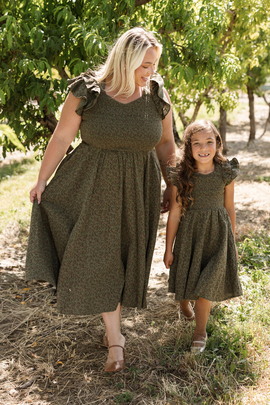 A woman and a young girl, both dressed in matching Ivy City Co Mini Hattie Dresses in the Woodland Floral print, walk together outdoors on a sunny day, surrounded by trees and grass.