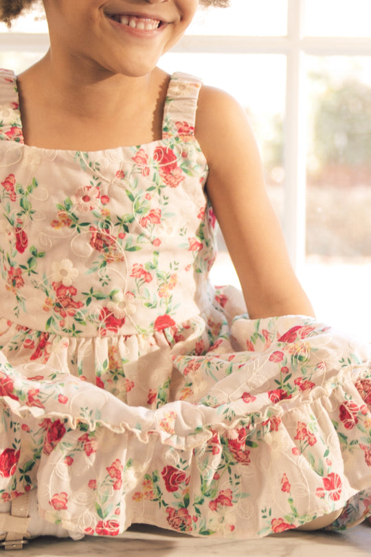 A smiling child sits in soft sunlight near a window, wearing the Ivy City Co Mini Helene Dress in Vibrant Chinz, with hands resting on their lap.