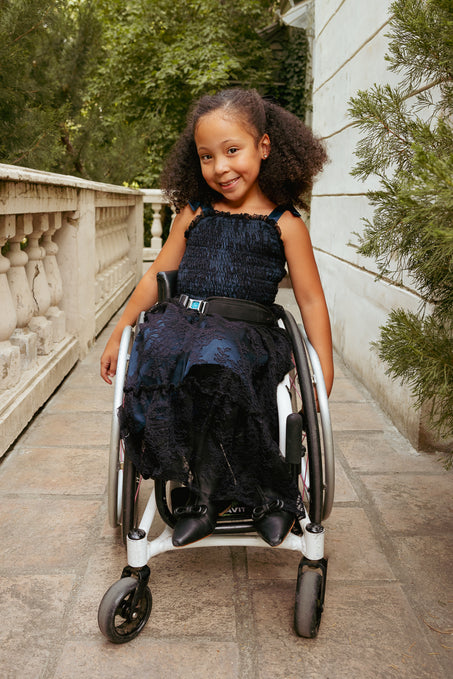Young girl in a wheelchair wearing a dark blue dress, sitting outdoors on a stone path with greenery.