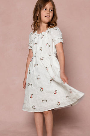 A young girl with light brown hair wears the Ivy City Co Mini Jessie Dress in Ditsy Embroidery, standing against a pink background and twirling as she looks slightly to the side.