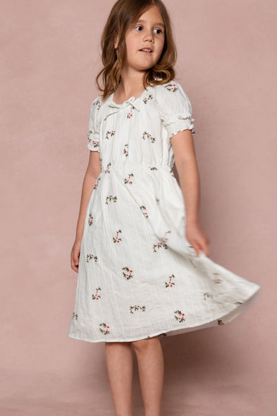 A young girl with light brown hair wears the Ivy City Co Mini Jessie Dress in Ditsy Embroidery, standing against a pink background and twirling as she looks slightly to the side.