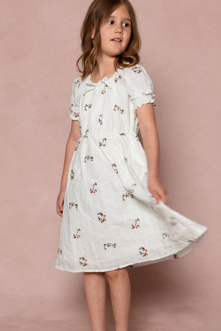A young girl with light brown hair wears the Ivy City Co Mini Jessie Dress in Ditsy Embroidery, standing against a pink background and twirling as she looks slightly to the side.