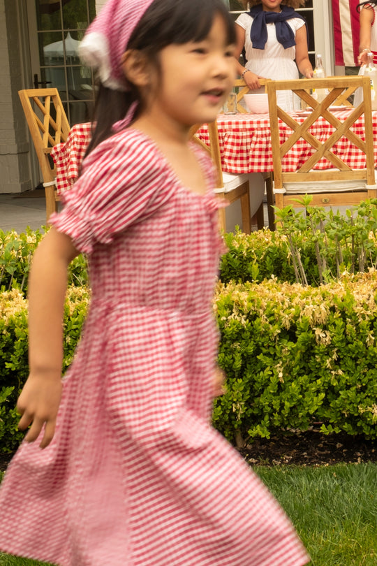 A girl wearing the Ivy City Co Mini Jessie Dress in Red Gingham runs by an outdoor table with a matching red gingham tablecloth; two adults and green hedges appear in the background, creating a festive Fourth of July scene.