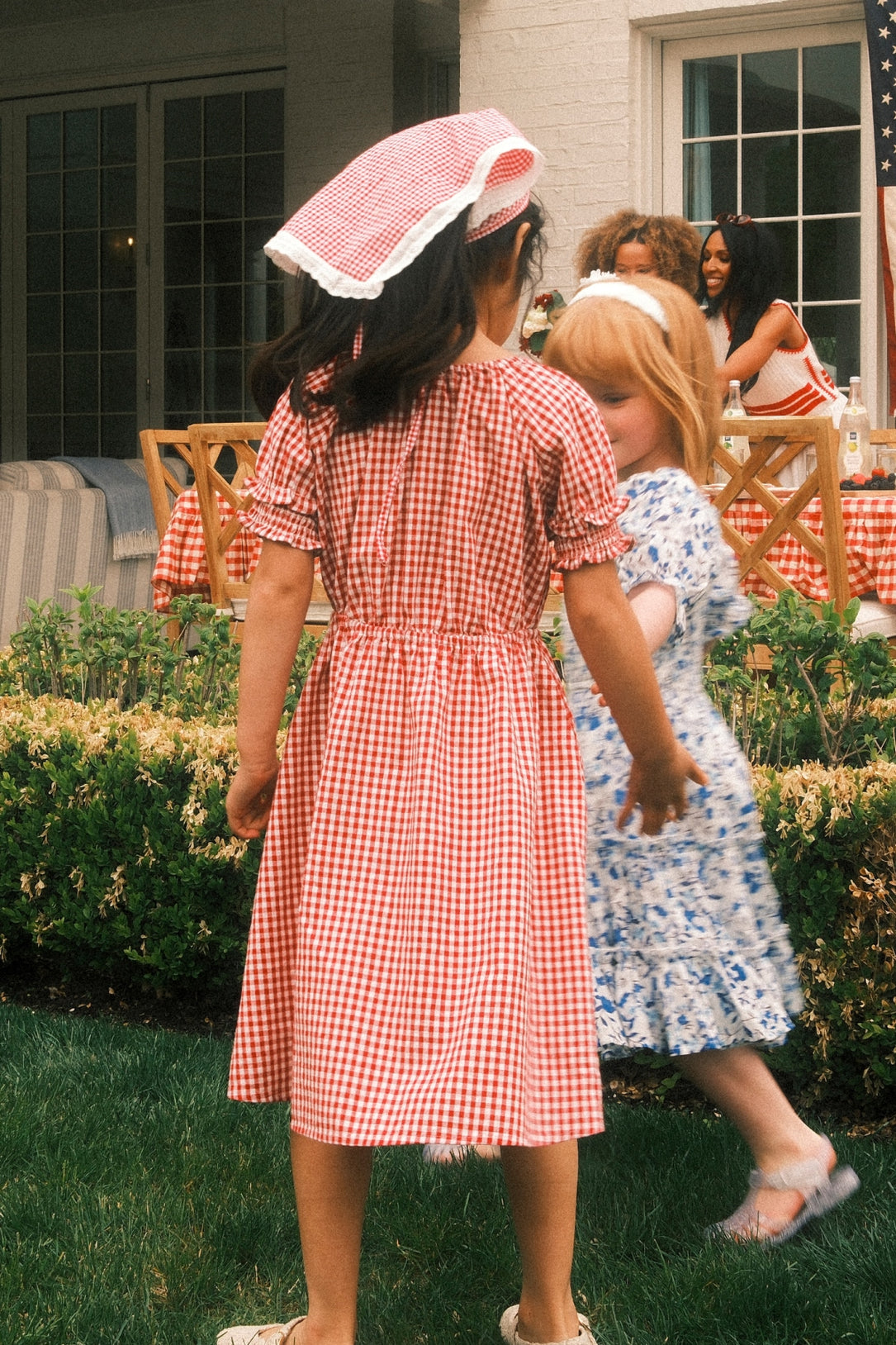 Two young girls play outside near a hedged garden. One wears the Ivy City Co Mini Jessie Dress in Red Gingham and a kerchief, while the other is in a blue floral dress. Adults sit at a table in the background, perhaps at a garden party.