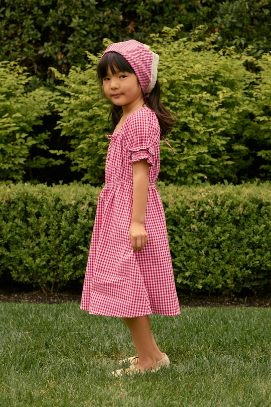 A young girl stands on grass in Ivy City Co's Mini Jessie Dress in Red Gingham—ideal as a girls’ Fourth of July dress—against a backdrop of lush green bushes and shrubs.