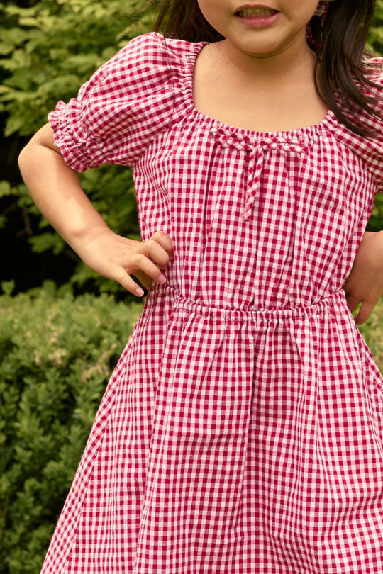 A child wearing the Ivy City Co Mini Jessie Dress in Red Gingham stands outdoors with hands on hips, surrounded by greenery. The classic red and white checks evoke a festive, timeless vibe; only the mouth and chin are visible in the image.