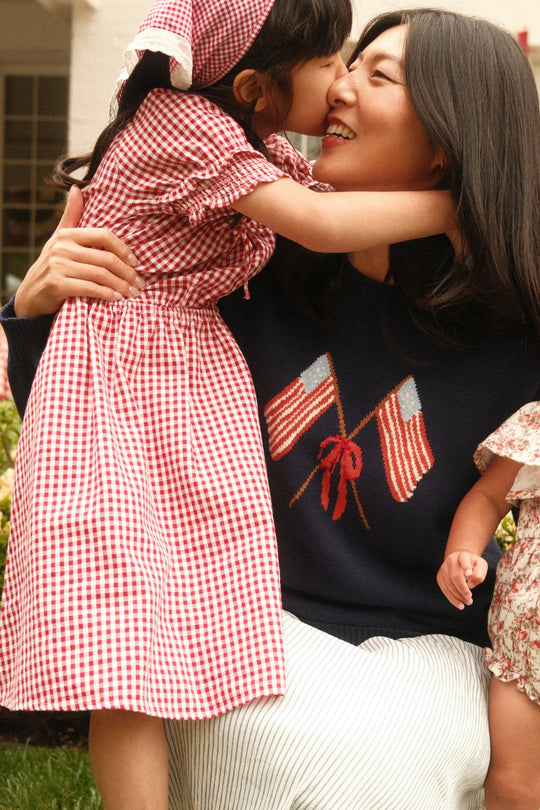 A woman smiles as a young girl in the Ivy City Co Mini Jessie Dress in Red Gingham and matching bonnet kisses her nose. Another child in a floral dress stands nearby. The woman wears a sweater with two flags, perfect for July 4th matching.
