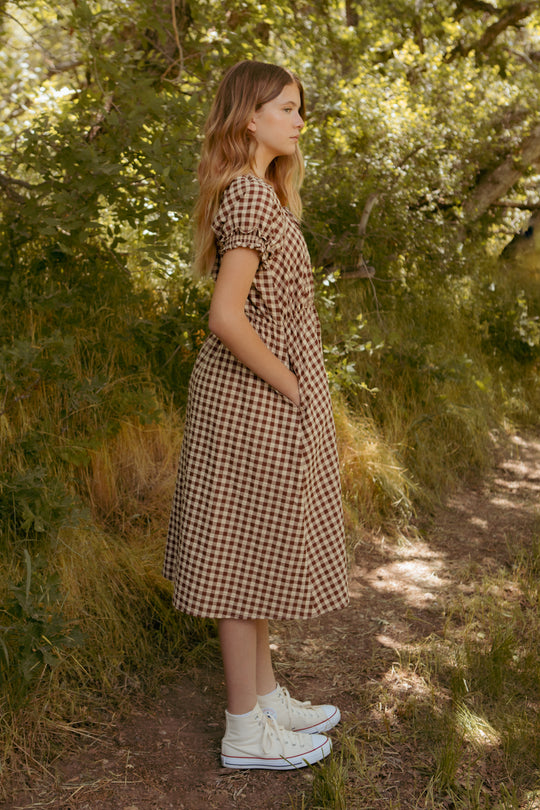 A young woman stands on a dirt path surrounded by greenery, wearing the Ivy City Co Mini Kaitlyn Dress in Chocolate Gingham with short sleeves and white high-top sneakers. She poses in profile with hands in her pockets among trees.