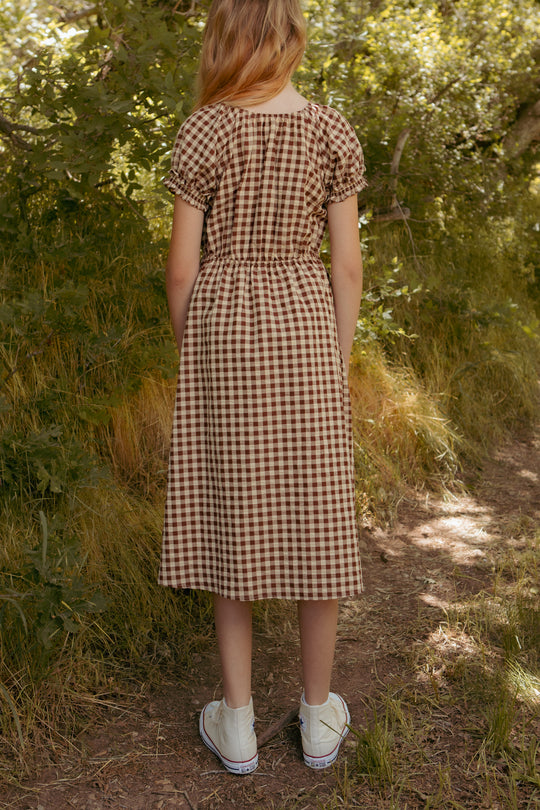 A girl with light brown hair, wearing the Ivy City Co Mini Kaitlyn Dress in Chocolate Gingham and white sneakers, stands on a sunlit dirt path in a green wooded area, facing away from the camera.