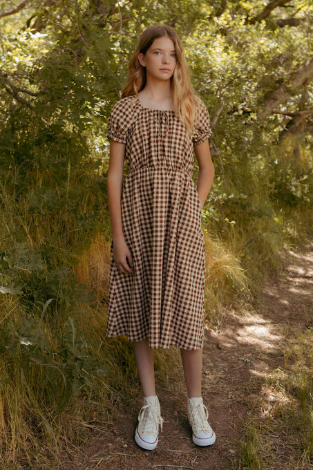 A young girl with long blonde hair stands on a dirt path, surrounded by green trees and grass. She wears the Mini Kaitlyn Dress in Chocolate Gingham by Ivy City Co, featuring a midi-length skirt, paired with white high-top sneakers.