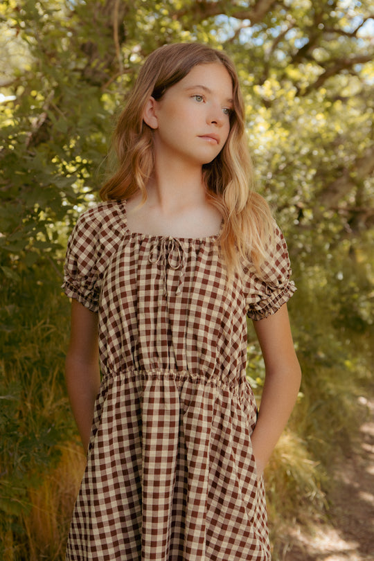 A young woman with long, wavy blonde hair stands outdoors among green trees, wearing the Ivy City Co Mini Kaitlyn Dress in Chocolate Gingham with short sleeves, looking to the side with a calm expression.