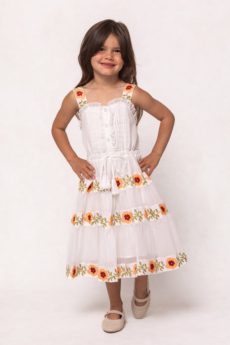 A young girl with long brown hair smiles, posing with hands on hips in the Ivy City Co Mini Katerina Dress in White with Floral Trim. The dress features colorful floral accents and the background is plain white.