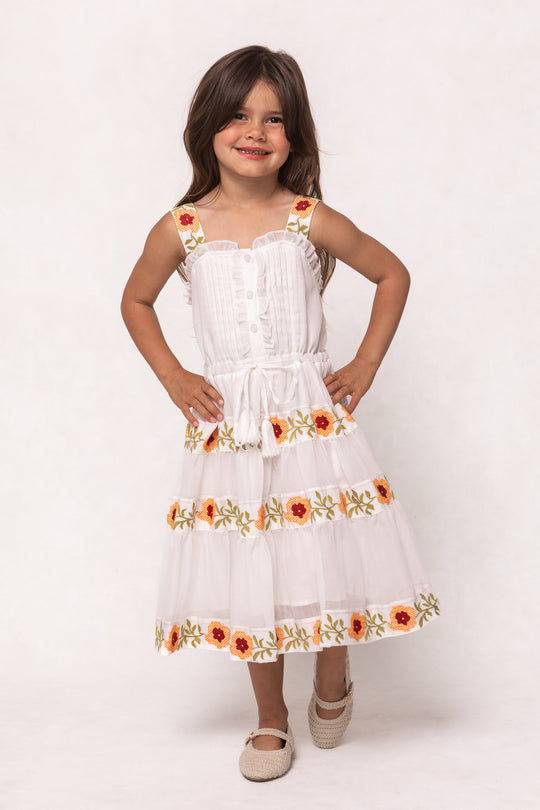 A young girl with long brown hair smiles, posing with hands on hips in the Ivy City Co Mini Katerina Dress in White with Floral Trim. The dress features colorful floral accents and the background is plain white.