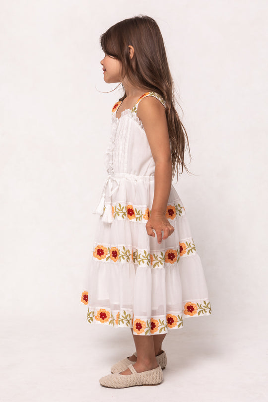 A young girl with long brown hair stands barefoot in profile against a plain background, wearing the Ivy City Co Mini Katerina Dress in White with Floral Trim—a sleeveless cotton voile dress with colorful embroidery and a matching belt.