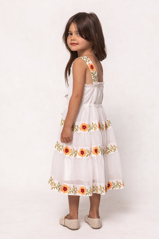 A young girl with long brown hair smiles over her shoulder in the Ivy City Co Mini Katerina Dress in White with Floral Trim, paired with beige woven shoes, set against a plain light background.