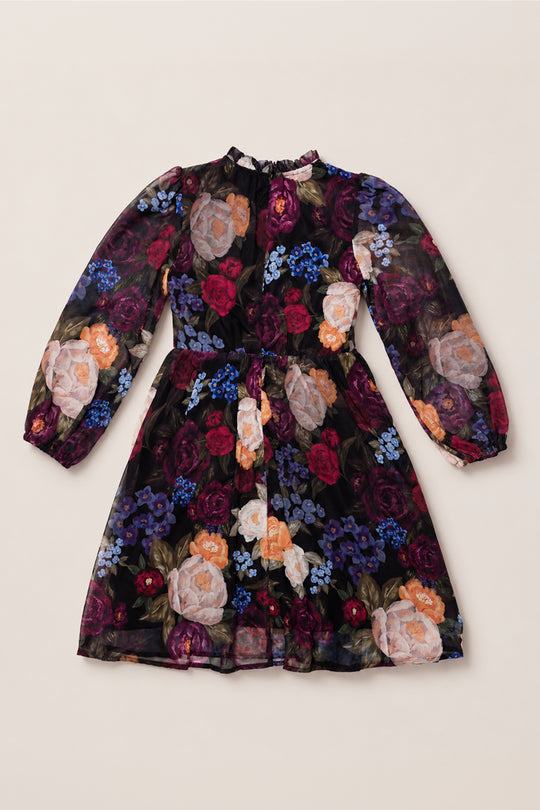 The Ivy City Co Mini Kelley Dress in Winter Floral features long sleeves, a knee-length silhouette, and a fitted waist. Its black background is adorned with a vibrant floral pattern in shades of red, purple, orange, and blue. Designed with a round neckline, this dress is perfect for holiday portraits or special seasonal gatherings.
