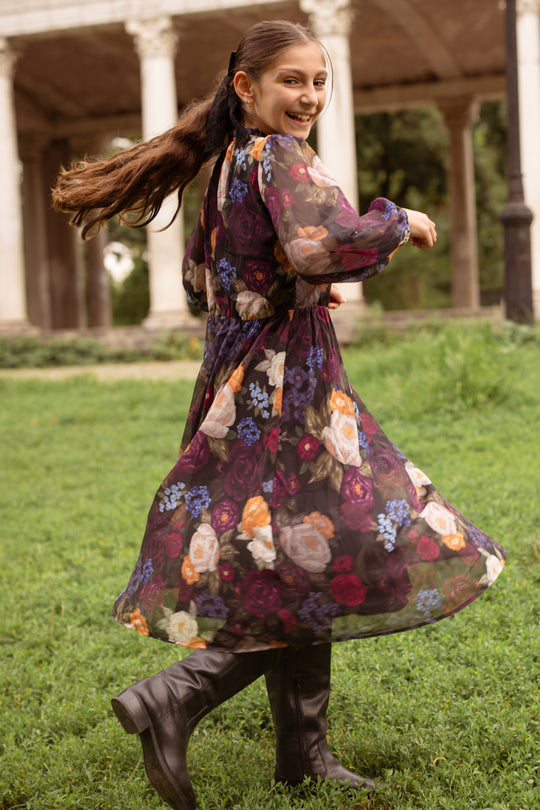 A smiling girl twirls on green grass in the Mini Kelley Dress in Winter Floral by Ivy City Co, paired with boots, with elegant columns and trees behind her—perfect for capturing holiday portraits.