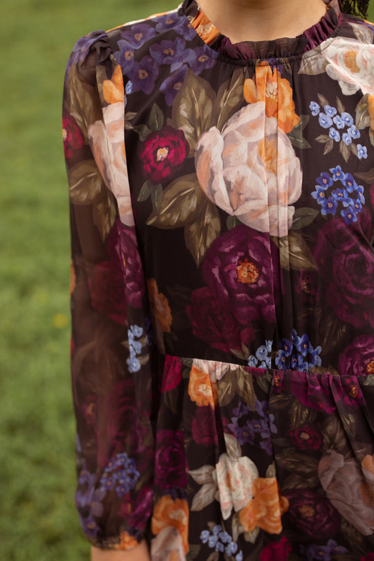 A person stands on grass wearing the Ivy City Co Mini Kelley Dress in Winter Floral, a dark, long-sleeved dress featuring a colorful floral pattern. The photo is cropped at the neck and mid-thigh, highlighting the details of this piece—perfect for holiday portraits.