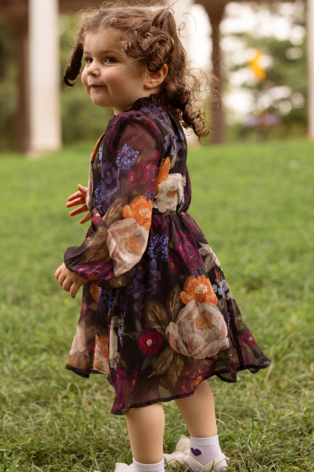 A young child with curly hair, wearing the Ivy City Co Mini Kelley Dress in Winter Floral, stands on green grass looking back over their shoulder and smiling. The blurred background—with its columns and greenery—creates a timeless setting for holiday portraits.