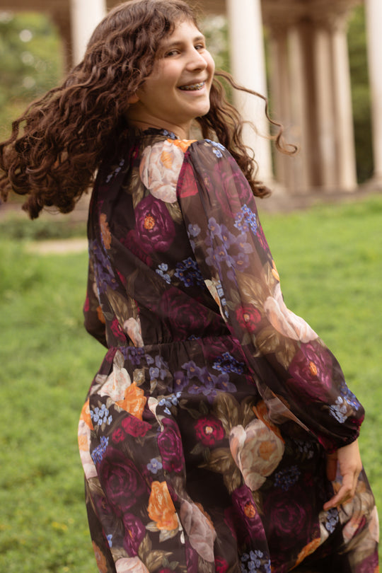 A woman with curly hair, wearing the Ivy City Co Mini Kelley Dress in Winter Floral, smiles while twirling outdoors. Green grass and stone columns in the background add to a charming setting, perfect for holiday portraits.