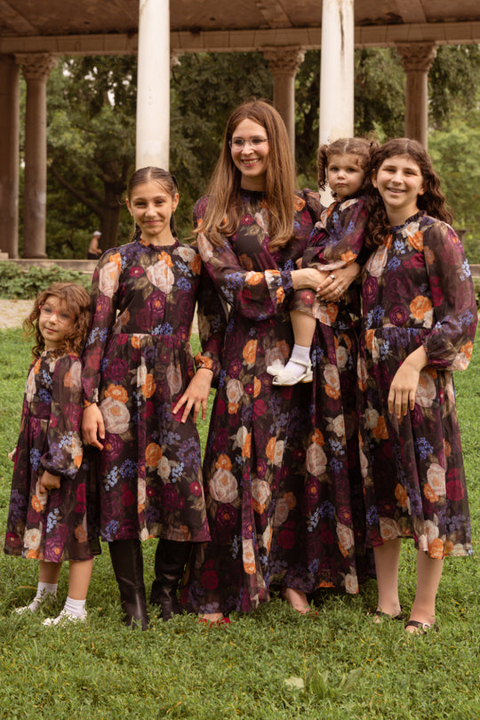 A woman and four young girls stand on the grass, all dressed in matching Ivy City Co Mini Kelley Dresses in Winter Floral—perfect for holiday portraits. The woman holds a toddler also outfitted in the charming Mini Kelley Dress as they smile together outdoors in front of tall columns and trees.