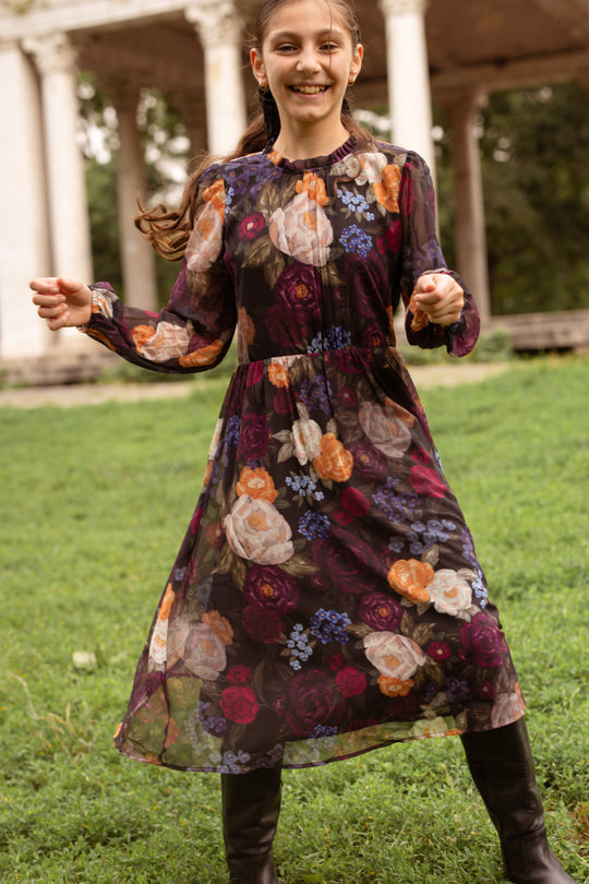 A smiling girl wears the Ivy City Co Mini Kelley Dress in Winter Floral with black boots, standing on green grass amid stone columns and trees—an elegant scene perfect for holiday portraits.