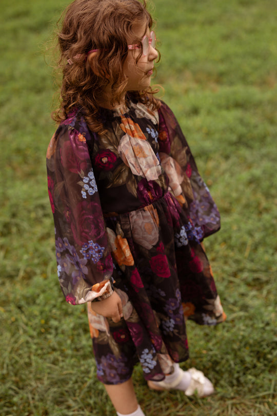 A young child with curly hair, wearing glasses and the Ivy City Co Mini Kelley Dress in Winter Floral, stands on green grass and looks to the side—perfect for charming holiday portraits.