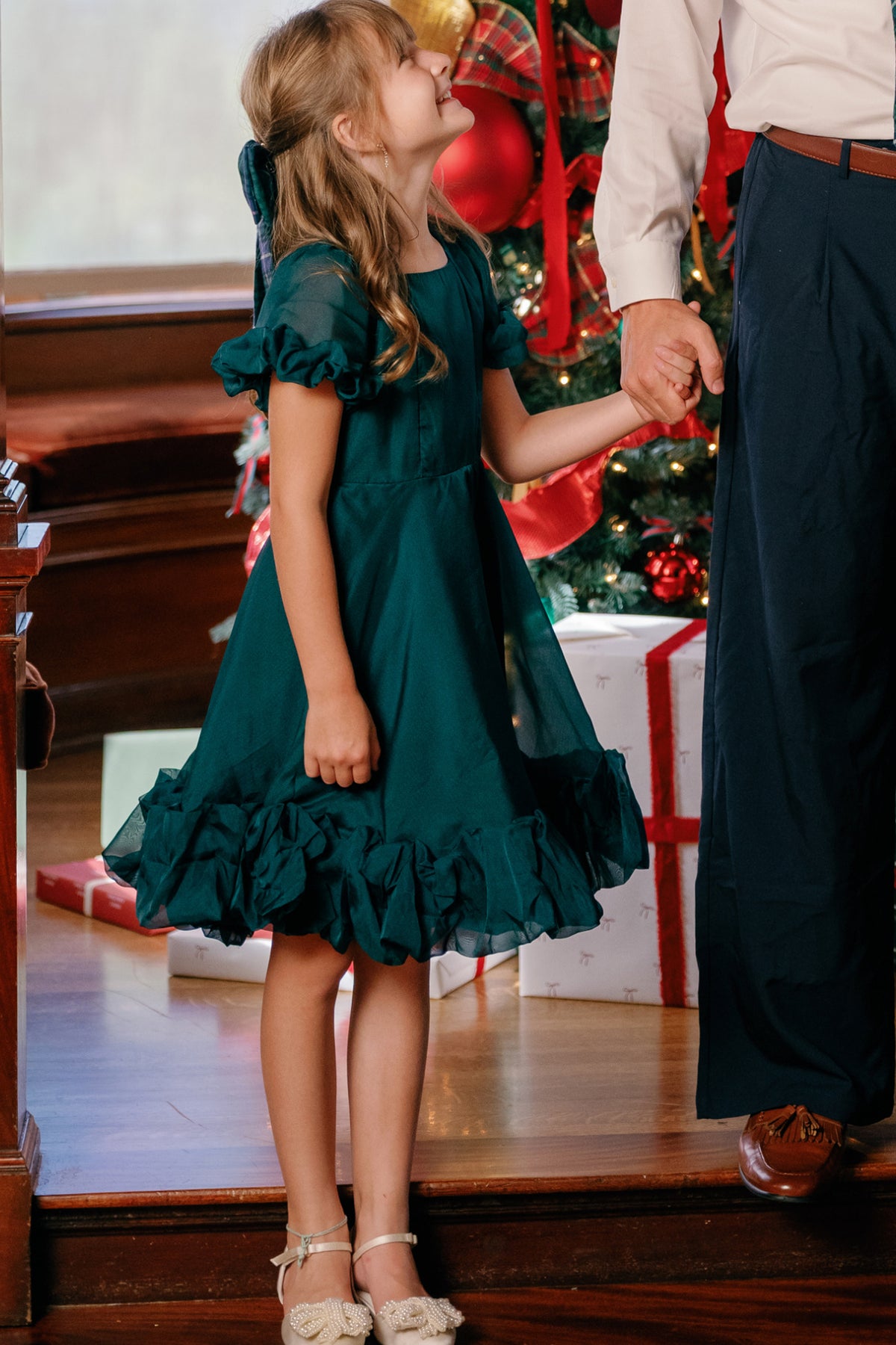 A young girl wears the Ivy City Co Mini Kendy Dress in Emerald, smiling as she holds an adult’s hand in front of a decorated Christmas tree and wrapped presents.