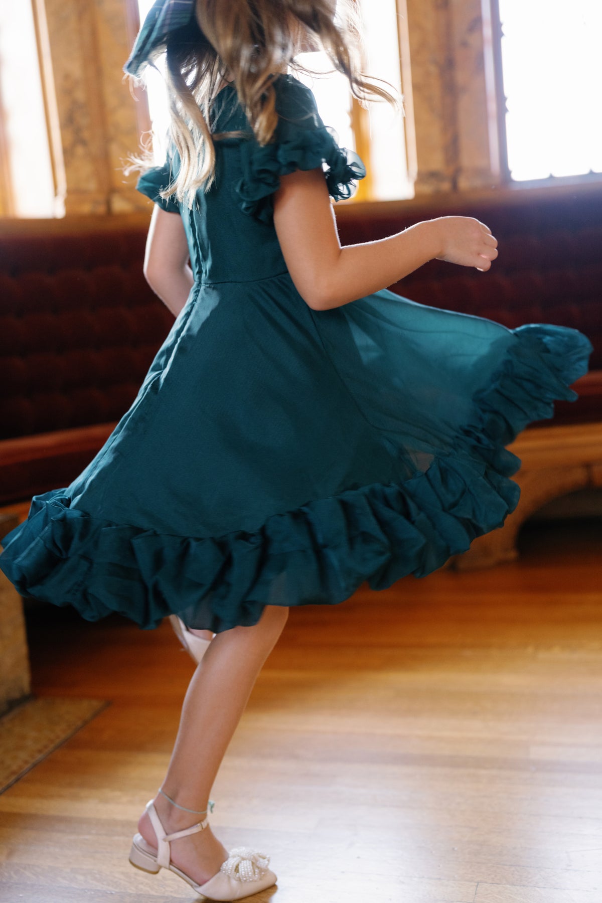 A girl twirls on a wooden floor in the Mini Kendy Dress in Emerald by Ivy City Co, sunlight streaming through the windows behind her.