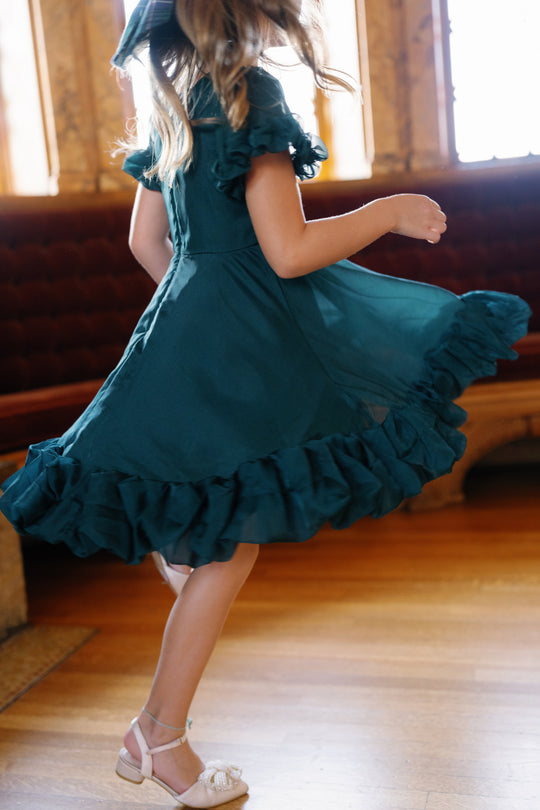 A girl twirls on a wooden floor in the Mini Kendy Dress in Emerald by Ivy City Co, sunlight streaming through the windows behind her.
