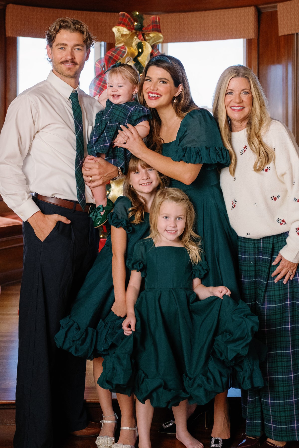 A family of five poses indoors by a decorated Christmas tree. The father wears a white shirt and plaid tie, while the mother and three daughters are dressed in matching Mini Kendy Dress in Emerald by Ivy City Co, perfect as girls holiday dresses. Another woman completes the festive scene in a white sweater and plaid pants.