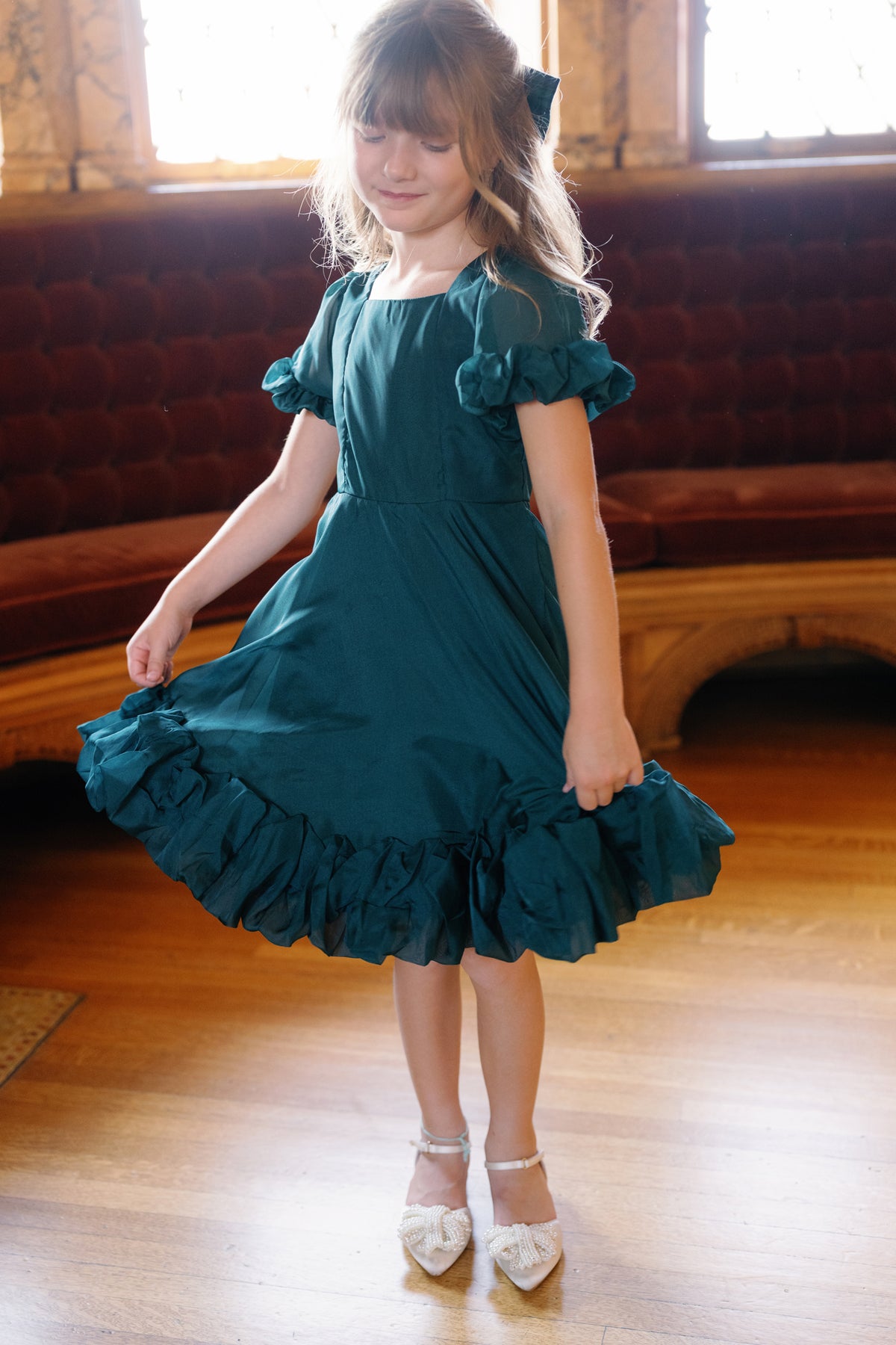 A young girl in the Ivy City Co Mini Kendy Dress in Emerald and white shoes stands on a wooden floor, gently holding her skirt. She is indoors near a velvet-cushioned bench and ornate windows.