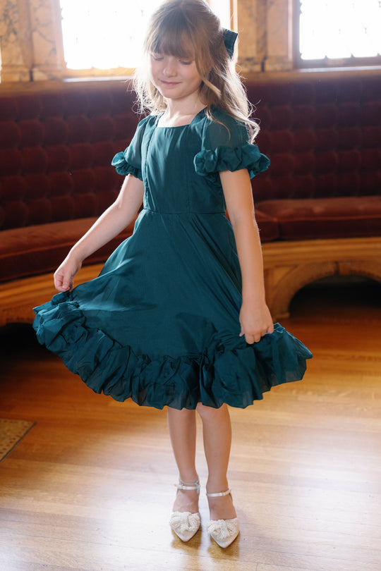 A young girl in the Ivy City Co Mini Kendy Dress in Emerald and white shoes stands on a wooden floor, gently holding her skirt. She is indoors near a velvet-cushioned bench and ornate windows.