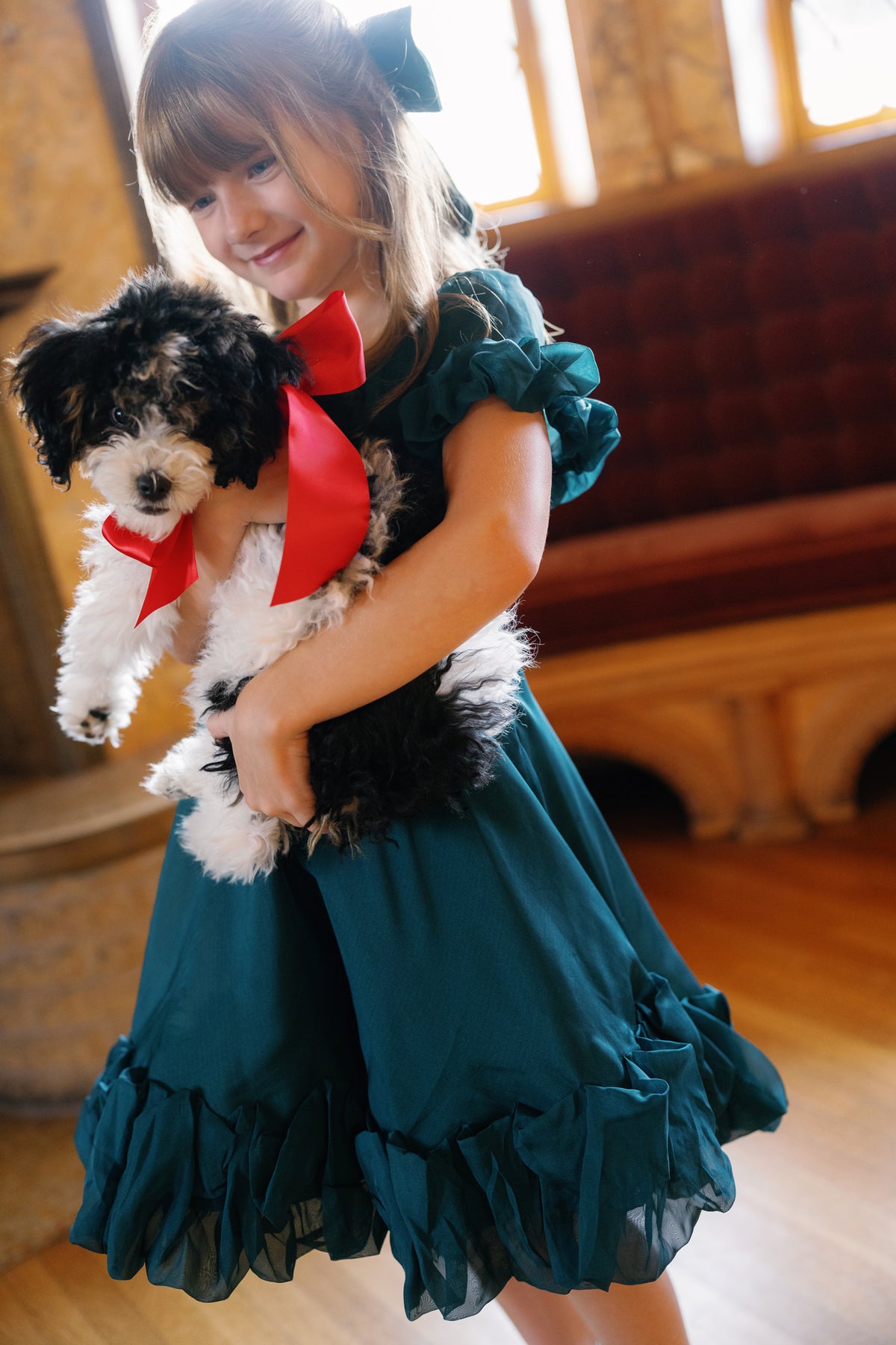 A young girl beams as she poses indoors in the Ivy City Co Mini Kendy Dress in Emerald, holding a small black and white dog adorned with a red bow. Sunlight streams through the windows, illuminating the cushioned bench behind them.