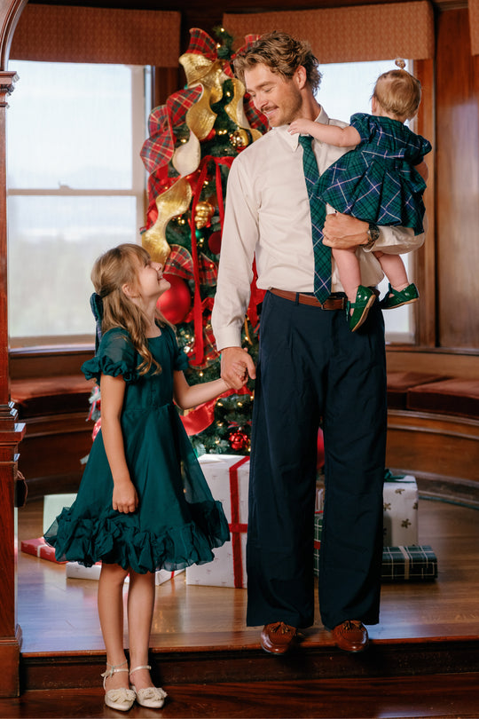 A man holding a toddler and holding hands with a young girl, all dressed up—the young girl in the Ivy City Co Mini Kendy Dress in Emerald—standing in front of a decorated Christmas tree with wrapped presents underneath.