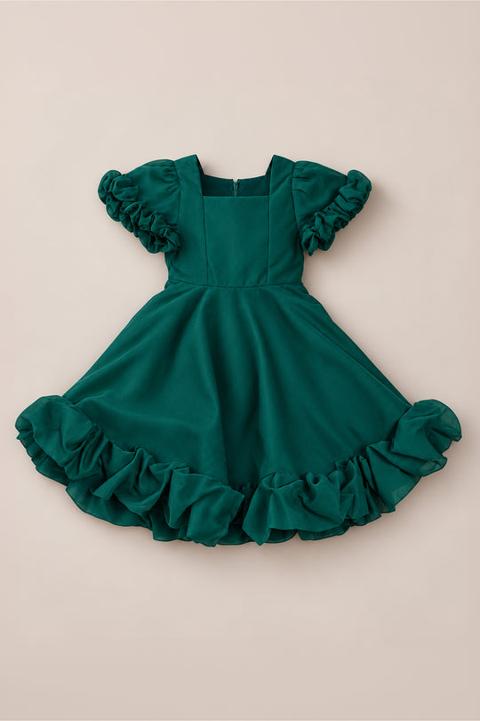 The Mini Kendy Dress in Emerald by Ivy City Co is a short-sleeved girls’ holiday dress with ruffled sleeves and a ruffled hem, shown laid flat against a light beige background.