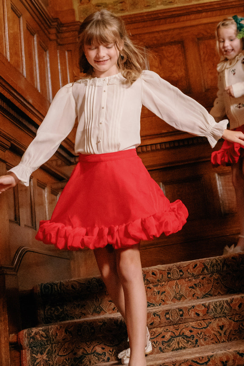 A young girl beams as she walks down carpeted stairs in a wood-paneled room, her holiday party look—the Ivy City Co Mini Kendy Skirt in Poinsettia paired with a crisp white blouse—bringing vibrant cheer to the space. Another girl dressed in a similar Ivy City Co outfit follows closely behind.