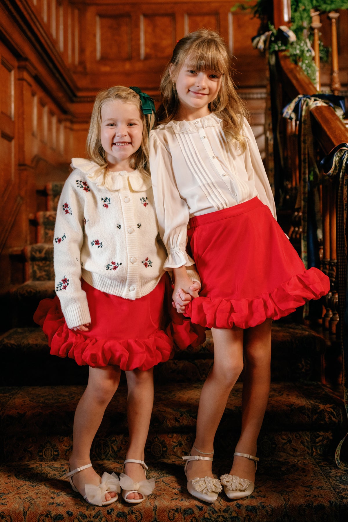Two young girls stand on a staircase, smiling and holding hands. Both are dressed for the holidays in Ivy City Co’s Mini Kendy Skirt in Poinsettia, paired with light tops featuring ruffled hems and dress shoes, surrounded by an elegant wooden interior.